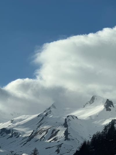 The image shows a partially snow-covered mountain range adjacent to a large cumulus cloud formation. A clear, blue sky is visible on the left side of the image. Dark green trees are present on the upper right side of the frame, along the mountain's slope. The scene appears to be photographed in daylight. The location is identified as Le Monêtier-les-Bains, France.