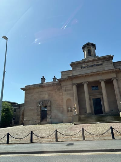 The image captures an upward angle of "The Courthouse" building in Knutsford, United Kingdom, under a clear blue sky. The building, made of light brown stone, features a classical architectural style with a prominent sign displaying "The Courthouse" and "Flat Cap Hotels". Its facade includes columns, a tower-like structure on top, and an ornate entrance. A chain fence runs along the perimeter, separating the building from the street. A streetlight is also visible.