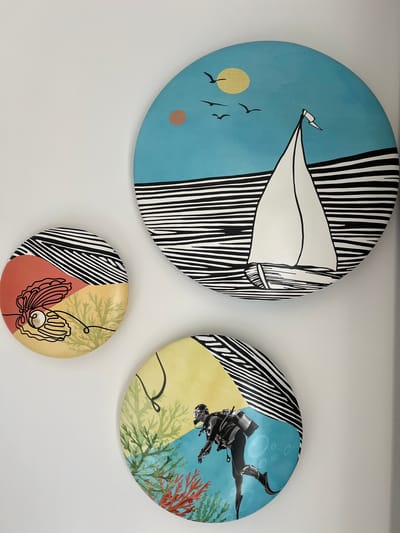 This image displays three circular artworks mounted on a white wall. The artworks feature stylized illustrations with a bold, graphic aesthetic.
The largest artwork, positioned on the left, depicts a light blue sky with a yellow sun and three silhouetted birds in flight. A white sailboat with a black and white striped sail dominates the upper portion of this plate. A small, tan circle is also present in the blue sky.
To the right of the largest artwork is a medium-sized plate. This piece features a diver in full gear swimming in a blue, underwater scene with air bubbles and colorful coral. The diver is depicted in a dynamic pose. The background transitions into a soft yellow area, intersected by the same black and white stripe motif seen on the other plates.
The smallest artwork, located below the other two, is divided into sections of color: black and white stripes, a reddish-brown, and a soft yellow. It features a line drawing of a shell with a pearl inside, and some green, sea-like vegetation.
The artworks collectively evoke a sense of adventure and the natural world, with elements of both sky and sea represented. The overall style is modern and visually striking. There is no visible text in the image. The location context is Ras Al-Khaimah, United Arab Emirates, but this is not explicitly indicated within the artwork itself.