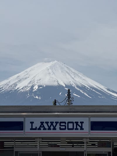 The image displays a serene yet striking landscape featuring the iconic Mount Fuji under an overcast sky. The snow-capped mountain, with its white slopes and visible patches of darker rock, occupies the majority of the background, stretching across the frame. Its peak is slightly softened by light clouds or atmospheric haze.
In the lower right foreground, a modern commercial building, identified as a Lawson convenience store, is visible. A prominent white sign with dark blue lettering clearly spells "LAWSON," framed by thin red and blue stripes. Parts of the building's facade, including dark-tinted windows and entryways, can be seen to the right of the sign, with a partially visible white sign showing "STOP" in red text on a door or gate.
A utility pole with multiple wires and attached equipment stands in the mid-ground, partially obscuring the lower slopes of Mount Fuji on the left side. A hint of green foliage is present at the very bottom right, suggesting nearby vegetation. The scene depicts a tranquil daytime setting, likely in Fujikawaguchiko, Japan, given the combination of Mount Fuji and the familiar Lawson store. No people or specific activities are visible, and the snow on the mountain suggests a cooler season.