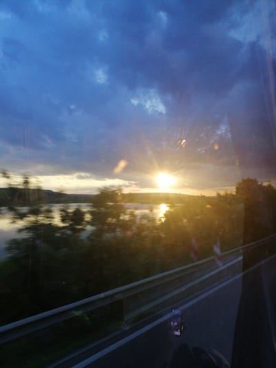 This is a photograph taken from inside a vehicle, likely a bus, looking out a window. The scene captures a sunset over a body of water with a landscape of hills and trees in the distance. The sky is dramatic with a mix of dark blue clouds and brighter areas where the sun is breaking through, creating a strong glare and lens flare effects on the window. The water reflects the colors of the sky and the surrounding environment. In the foreground, a highway guardrail is visible, running parallel to the road. The overall impression is of a journey at dusk, with the natural scenery being the primary focus. The location is identified as Pocoleni, Romania, suggesting the photograph was taken in or near this area. There are no people clearly visible in the frame.