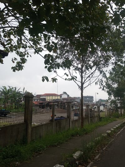This image captures an outdoor scene in Houaythat, Laos, viewed under an overcast sky. The upper foreground is obscured by dense, dark green tree foliage. A concrete sidewalk, partially covered by green grass and weeds, runs along the lower right, parallel to an asphalt road separated by a segmented concrete curb. Across the midground, a concrete wall topped with a metal fence and regularly spaced concrete posts forms a boundary. Behind this barrier, to the left, is an unpaved area containing piles of dark aggregate material and the partial view of a black utility trailer or truck bed. Further left, a stand of tall banana trees is visible. To the right of the fence, a white panel van is parked on a grassy patch. The background consists of a collection of buildings, including a multi-story structure with a red roof and pale yellow facade, and various other grey and white buildings. A street lamp is visible on a pole, and a slender communication tower rises in the distant background. No individuals are clearly discernible.