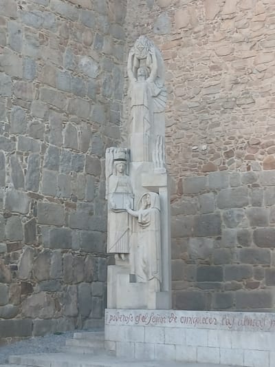 The image shows a stone sculpture in front of a stone wall in the city of Avila, Spain. The sculpture is a tall, slender monument composed of several figures. At the top, a large figure with outstretched arms appears to be holding a circular object or crown, with wings or decorative elements extending from its sides. Below this figure, a smaller figure stands with a headdress and holds an object that resembles a book or tablet. To the right of this figure, a third, smaller figure, draped in robes, extends a hand towards the second figure. The base of the sculpture has red lettering carved into it. The stone wall behind the sculpture is constructed from rough-hewn, irregular stones of varying sizes and colors, with visible mortar lines. The ground in front of the sculpture consists of a small set of stone steps leading up to the monument, with gravel and some sparse vegetation around the steps. The lighting suggests it is daytime, with the sun casting some subtle shadows.