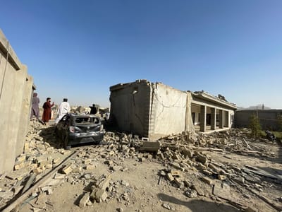 The image depicts a scene of extensive structural damage in Spin Boldak, Afghanistan, under a clear, bright blue sky. A dark-colored, severely damaged sedan, missing its rear window and exhibiting significant body distortion, rests amidst a large accumulation of concrete and brick rubble in the foreground.
To the left, a partial concrete wall stands damaged. To the right, a single-story building with a porch supported by columns is in a state of partial collapse; its exterior wall has fallen, exposing internal structures and creating a large debris field extending into the foreground. Cracks are visible on the remaining wall surfaces.
Several adult males are present. One man in a white tunic and another in a brown tunic stand near the damaged vehicle on the left, observing the scene. Additional figures are visible further back along a debris-strewn path. In the far right background, a motorcycle is parked near a small tree, and a tall, slender structure, possibly a minaret, is visible on the horizon. The ground is dry, dusty earth covered extensively with debris from the collapsed structures.