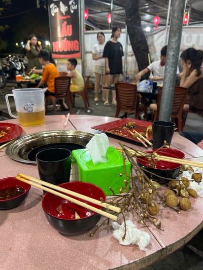 The image depicts a street-side dining scene, likely in Minh Xuan Ward, Vietnam, at night. A pink-tiled, circular table is laden with remnants of a meal. In the foreground, two red and black bowls sit with chopsticks resting on them. One bowl contains a dark dipping sauce, while the other is mostly empty with some scattered food debris. To the left, a large, clear plastic pitcher filled with a golden liquid, possibly beer, is present. Next to it, a metallic pot with a black interior, likely for cooking, is visible. A green tissue box is positioned centrally. Scattered around the table are several longans, some still on the branch, and crumpled white tissues. In the background, people are seated at tables, some eating and others engaged in conversation. A large sign with Vietnamese text, likely advertising "LẨU NƯỚNG" (hot pot and barbecue), is prominently displayed. The environment is illuminated by ambient light from street lamps and overhead fixtures, creating a casual, lively atmosphere.