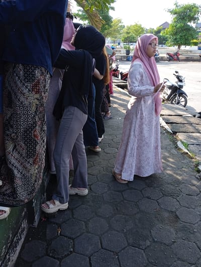 The image shows a line of people in Kauman, Indonesia. Most of the people are wearing hijabs and casual clothes. A woman wearing a light pink hijab and a matching long dress is standing slightly apart from the line, holding a piece of paper. Motorcycles are parked nearby. The ground is paved with grey hexagonal blocks.