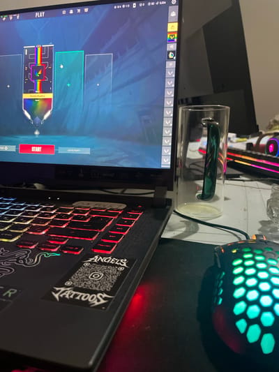 A laptop displays a video game menu with "PLAY" and "START" prominently visible. The game features a map with colorful representations. A glass mug with a green handle rests near the laptop. Adjacent to the laptop is a gaming mouse with a honeycomb pattern and green lighting. The keyboard of the laptop has red illuminated keys and stickers. The environment includes elements of a gaming setup, such as a mousepad and potentially other gaming peripherals in the background. The photo does not show the location in the city of Oued Ellil, Tunisia.