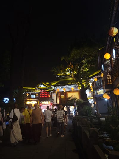 A nighttime outdoor scene depicts a pedestrian street in Wuhou District, China, featuring traditional Chinese architecture adorned with yellow and red lanterns. Several individuals are walking along a paved pathway. In the foreground, three people are walking together: a person in a white outfit with a black bag, a person in a bright yellow shirt and dark pants, and a person in a brown tank top and dark pants. To the right, the street is lined with multi-story buildings with dark wood facades and decorative lighting. Numerous yellow lanterns hang from the buildings and trees. A decorative railing runs along the right side of the path, bordering a small water feature. The overall ambiance is that of a bustling night market or cultural street.
