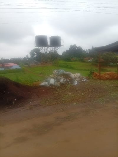 Two large black water tanks are elevated on a metal structure in a rural landscape under a cloudy sky. Green fields and trees stretch into the distance with scattered buildings. A pile of white bags and debris sits on the ground near a dark mound. In the foreground, a muddy path or road is visible. A shed-like structure is on the right edge of the frame. Overhead power lines traverse the sky.