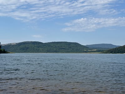 This image depicts a serene, natural landscape featuring a wide, calm body of water in the foreground and rolling, tree-covered hills in the background. The sky above is a clear, pale blue with scattered, wispy white clouds. The water's surface is slightly rippled, reflecting the soft light of the day. The hills, densely forested with green foliage, rise gradually, with some showing rocky outcrops along their crests. The overall impression is one of tranquility and natural beauty.
The scene appears to be set during the daytime, with the sun casting a gentle light that suggests it is either morning or late afternoon. The weather seems pleasant and mild, indicated by the clear sky and calm water. There are no people or man-made objects prominently visible, emphasizing the unspoiled nature of the location. The landscape suggests a rural or wilderness setting, possibly a lake or a reservoir. The absence of any visible text or specific identifying landmarks makes it difficult to pinpoint an exact location beyond the general type of natural environment. The mood evoked is peaceful and contemplative, inviting the viewer to appreciate the quiet majesty of the natural world.