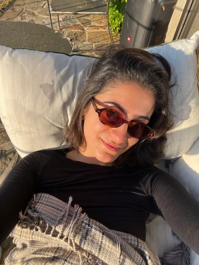 A woman is lying on a white pillow outdoors in Gondarém, Portugal. She is wearing sunglasses and a black long-sleeved shirt and is partially covered by a fringed blanket. A black speaker is visible on the left of the frame, and the area around the pillow is paved with stones. The scene appears to be lit by direct sunlight.