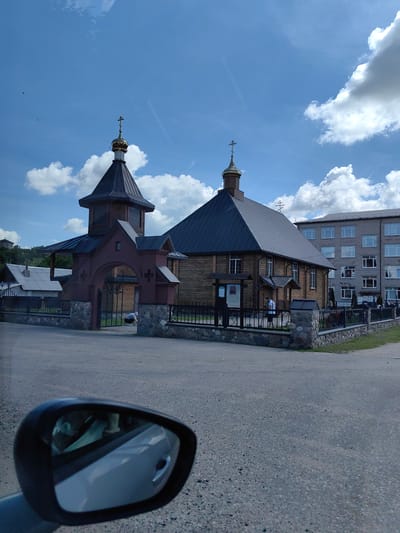In the city of Ilyanskiy selskiy Sovet, Belarus, a scene unfolds under a bright, cloudy sky. A traditional wooden Orthodox church with a dark metal roof and a golden dome stands prominently in the frame. The church features a bell tower topped with a cross and ornate windows. A red brick gateway with stone accents and metal fencing surrounds the church grounds. To the right of the church, a multi-story grey building with numerous windows is visible. In the foreground, a car's side mirror reflects a partial view of the interior of the vehicle, including what appears to be a person with a face mask. The ground is paved with asphalt, and a patch of green grass is visible behind the fence.
