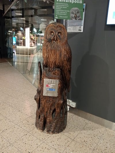 The image shows a carved wooden statue of an owl perched on a tree stump, situated indoors in a public space with a speckled tiled floor and glass walls revealing shops in the background. The owl statue is brown and intricately carved, with large eyes and detailed feathers. A metal plate is affixed to the tree stump, featuring a slot and some stickers, one of which appears to be a QR code. To the right of the statue, electrical outlets are visible on a dark gray wall. A green poster with text and an image of an owl is mounted on the wall above the statue. Based on the presence of the poster in Finnish, the location is likely in Heinola, Finland. The scene is well-lit and appears to be a commercial or public area. There are no people visible in the image.