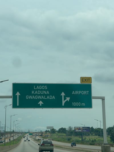 An eye-level, medium shot captures a green highway sign with white lettering and arrows, indicating destinations and distances. The sign reads "LAGOS KADUNA GWAGWALADA" with an upward arrow on the left, and "AIRPORT" with a right-turn arrow and "1000 m" on the right. Above the green sign, a yellow rectangular sign reads "EXIT." The sign is positioned over a multi-lane highway with vehicles in motion. The sky is overcast with grey clouds. To the right of the green sign, a billboard is visible. The highway is bordered by grassy verges and trees. The scene is located on a highway leading towards the Immigration Quarters in Nigeria.