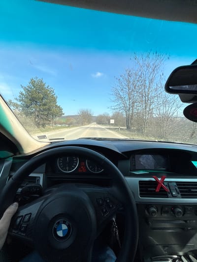 The image displays a first-person perspective from the driver's seat of a black BMW E60 series vehicle, traveling on a paved rural road in Erden, Bulgaria. A left hand, wearing a blue sleeve, is visible gripping the multi-function steering wheel. The instrument cluster shows a speedometer indicating approximately 40 km/h, a tachometer, and a digital display reading 11:54, an outside temperature of 6.4°C, and vehicle information. The center console features an infotainment screen displaying a grey, textured image. A red 'X' shaped accessory is clipped into the rightmost air vent. Vehicle keys are visible hanging from the ignition. The windshield appears dusty with some smudges.

Outside the vehicle, a relatively straight, two-lane asphalt road extends into the distance under a clear blue sky with minimal cloud cover. To the left, a prominent evergreen pine tree stands beside a grassy area, with several white, rectangular structures, possibly beehives, further in the background. Beyond these, green fields and rolling hills are visible. To the right, numerous leafless deciduous trees line the roadside, consistent with late winter or early spring conditions. A small white sign with red elements is positioned on the right shoulder of the road, its text unreadable.