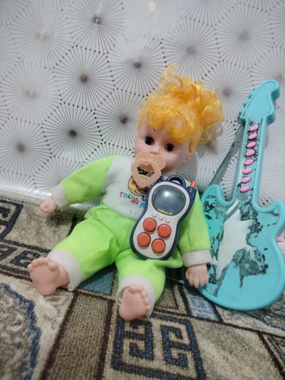 In Cholpon-Ata, Kyrgyzstan, a doll with blonde hair and a pacifier rests on a rug, adorned in a green outfit. A toy cell phone is placed on its chest, and a blue toy guitar lies beside it. The background features a patterned wallpaper.
