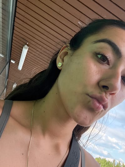 The image presents a close-up, low-angle portrait of a young adult female. She has long, dark hair, dark brown eyes, and fair skin with visible pores and several small moles on her neck and chest. Her lips are puckered into a pout, and her gaze is directed upwards and to her right. She is wearing a gray sleeveless top, a thin gold-colored chain necklace, and a white and yellow daisy-shaped stud earring in her visible right ear.
The upper background features a structure composed of parallel brown wooden planks, suggestive of a ceiling or overhang. A single, elongated white fluorescent-style light fixture is mounted within this wooden structure. The lower background transitions to an outdoor scene, revealing a blue sky with scattered white cumulus clouds and a distant horizon line of green vegetation, consistent with trees or bushes.
There are no discernible visual elements within the image that specifically identify the location as Imatra, Finland.