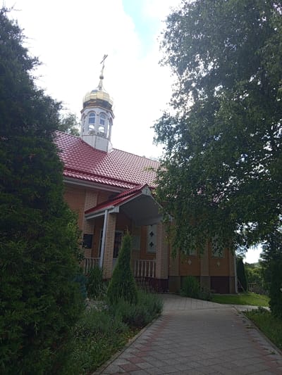 This image depicts an Orthodox church in Giaginskaya, Russia. The church is constructed of light-colored brick with a red tiled roof. It features a golden dome topped with a cross, and a white bell tower with arched windows. A paved walkway leads to the entrance, which is covered by a red awning. Evergreen trees and other greenery surround the church. The sky is partly cloudy.