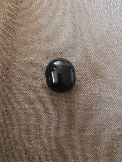 A black, oval-shaped charging case for wireless earbuds rests on a textured, brown fabric surface. The case is glossy and reflects light, highlighting its smooth, rounded form. The fabric has a coarse weave and appears to be a common upholstery material. There are no people, animals, or other objects visible in the image. The scene is stationary and devoid of any action. The image does not contain any information that would allow for its location in Dranda, Abkhazia to be determined.
