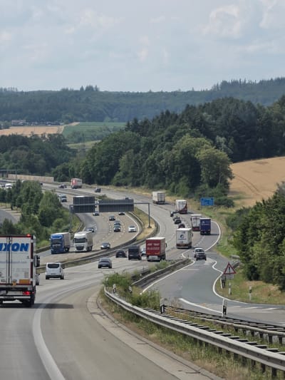 The image captures a multi-lane highway scene on a slightly overcast day, likely near Montabaur, Germany, given the presence of a German highway sign. The highway curves to the right, with vehicles traveling in both directions. A large white truck with "DIXON INTERNATIONAL LOGISTICS" branding is prominently featured in the foreground on the left. Several other trucks and cars are visible on the highway, including a blue truck, a white truck, and various passenger vehicles. The surrounding landscape consists of dense green trees and fields of golden crops, suggesting a rural or semi-rural setting. The sky is filled with scattered clouds. The highway infrastructure, including guardrails and signage, is clearly visible.