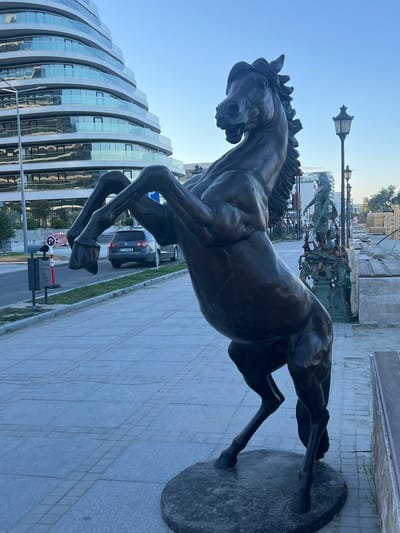 A bronze statue of a rearing horse stands on a stone base on a paved walkway. The horse is dark brown and detailed, with its front legs raised in the air and its head turned to the left. In the background, a modern building with curved glass balconies is visible. To the right of the statue, there is a street with a car and a traffic sign. Further in the background, there is a lamp post and another statue of a person. The scene appears to be in a public area in the city of Năvodari, Romania, likely a park or promenade, with construction materials visible in the far background. The sky is clear and blue, suggesting it is daytime.