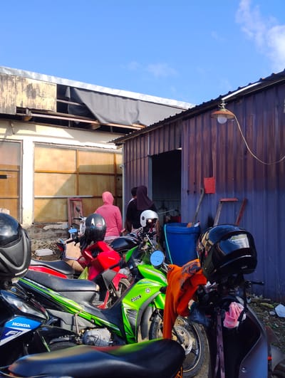 A bright, sunny day with a clear blue sky is depicted in Pompanua, Indonesia. The scene focuses on a cluster of parked motorcycles in front of a weathered, corrugated metal building. The building has a rustic appearance with visible rust streaks on its exterior. Adjacent to it is a lighter-colored structure with large, boarded-up window openings, suggesting it might be under construction or renovation.

Several people are visible, mostly standing near the entrance of the metal building. One person wears a bright pink hooded jacket, another a dark headscarf, and a third is partially obscured.

The foreground is dominated by several motorcycles. A bright red motorcycle is prominent, with a black helmet resting on its handlebars. Next to it, a green and black motorcycle has an orange garment draped over its handlebars, with another helmet on top. Another black motorcycle with blue "HONDA" branding is partially visible to the left. The ground around the motorcycles is rough, with some debris and a large blue plastic barrel. The overall impression is of an outdoor area associated with a functional, possibly industrial or workshop space.