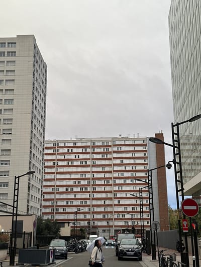 The image displays an urban street scene in Levallois-Perret, France, under an overcast sky. Three distinct multi-story buildings are visible: a tall, light-grey residential tower on the left, a wider residential building in the center-background characterized by horizontal bands of reddish-brown brickwork alternating with rows of white-framed windows, and a modern, glass-clad high-rise on the far right.
The street is lined with multiple dark-colored streetlights, some of which are equipped with surveillance cameras. A dense cluster of motorcycles and scooters is parked on the left side of the street. Further along, a dark grey hatchback and a black sedan are parked, with another vehicle displaying illuminated headlights visible in the distance. A red circular 'no entry' sign is positioned on the right side of the street.
In the foreground, slightly right of center, a young woman walks to the right, holding a transparent umbrella over her left shoulder. She is dressed in a light-colored jacket and dark trousers, and carries a dark shoulder bag.