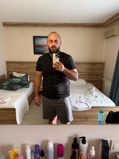 This is a mirror selfie taken in what appears to be a hotel room in Ayvalık, Turkey. The photo is oriented vertically. The primary subject is a bald man with a beard, wearing a black t-shirt with a white anchor pattern and gray shorts. He is holding a smartphone in his left hand, which is used to take the photo, and his right arm is visible at his side.

The setting includes a bed with white linens and pillows, a wooden headboard, and a framed picture on the wall depicting a sunset over water. In the background, reflected in the mirror, is a wall with an air conditioning unit and a wooden paneled closet or wall. To the right of the mirror, a shelf unit holds several bottles and containers, likely toiletries, including what appears to be a bottle of water and various personal care products. One bottle has "Sun Dance" printed on it. The lighting suggests it is daytime. The overall mood is casual, possibly the individual is getting ready or has just woken up.