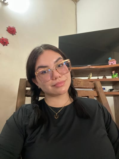 A young woman with dark hair in pigtails and large, transparent-rimmed glasses is seated in a wooden chair, looking towards the camera. She is wearing a black long-sleeved shirt and a gold necklace with a heart-shaped pendant. In the background, a wooden shelf unit is visible, displaying various decorative items, including small figurines and a television screen. Ladybug decorations are affixed to the wall to the left of the woman. The image appears to have been taken indoors, possibly in a residential setting. There is no specific indication of the location being in San Antonio, Venezuela.