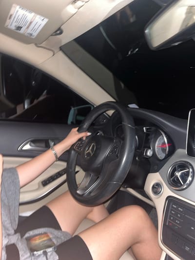 A person is seated in the driver's seat of a Mercedes-Benz car. The person is wearing grey clothing and black thigh-high socks. The person's left hand is on the steering wheel, with a silver bracelet visible on the left wrist. The car's dashboard, steering wheel, and interior are visible. The background outside the car windows is dark. The location is not possible to determine based on the image.