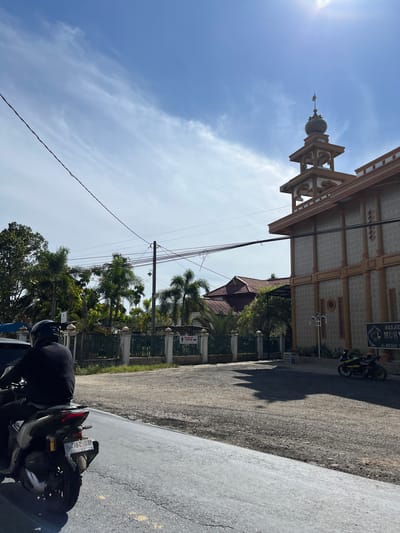 The image shows a street scene in Bukit Rata, Indonesia, on a bright, sunny day with a few scattered clouds. The primary subjects are a mosque with a distinctive minaret and dome, a street with a gravel shoulder, and a motorcycle.

The mosque's architecture is visible from the side, with its patterned facade and decorative elements. The minaret, topped with a dome, stands prominently against the blue sky. Palm trees and other lush greenery surround the mosque, suggesting a tropical or subtropical location. A white fence encloses a section of the mosque's grounds.

A street runs alongside the mosque, with a dark asphalt surface and a lighter, lighter-colored road to the right. A motorcycle, parked on the right side of the road, is partially visible in the foreground. A person wearing a dark shirt and helmet is seated on the motorcycle, facing away from the camera.

Visible text on a sign above the mosque reads "MASJID MUH...", with "JL. MEDAN..." partially visible below it. Another sign with an arrow pointing to the right reads "WUDUK PRIA" which translates to "Men's Ablution Area".

The bright sunlight casts strong shadows, indicating it is likely midday or early afternoon. The overall atmosphere is peaceful and everyday, depicting a common scene in a community with a prominent place of worship.