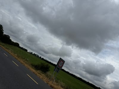 The image depicts a scene on a road, likely in Kilivory, Ireland, given the context. The road is paved with asphalt and has yellow dashed lines indicating lanes. The sky is heavily clouded with dark, dramatic storm clouds. To the right of the road, a speed limit sign is visible, showing "100 km/h" within a red circle, mounted on two metal poles. Beyond the road and the sign, a vast expanse of green farmland stretches towards a line of trees in the distance. The land is a vibrant green, suggesting it is well-maintained pasture or fields. The overall atmosphere is overcast and somewhat ominous due to the thick cloud cover. There are no people or animals visible in the frame. The composition is at an angle, giving a sense of movement or perspective from a vehicle traveling along the road.