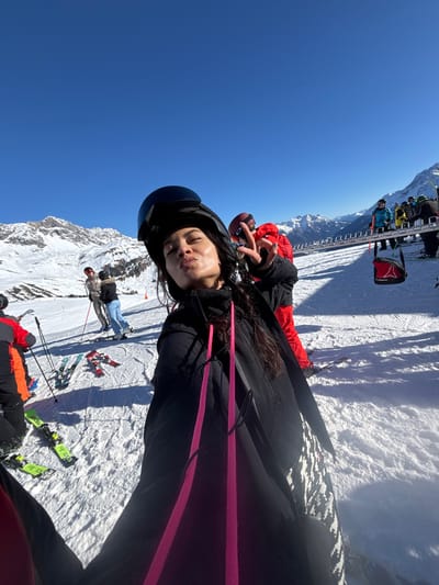 This image captures a bright, sunny day on a snow-covered mountain slope in St. Christoph, Austria.
In the foreground, a female individual is seen taking a selfie, positioned close to the camera. She wears a black ski helmet and a black ski jacket with prominent neon pink drawstrings. Her dark hair is visible beneath the helmet. Her face is turned slightly upward, eyes closed, with lips pursed. Her left hand is raised, forming a "peace" sign. Directly behind her and to the right, another individual in a red ski jacket, dark helmet, and goggles also displays a "peace" sign with their right hand.
In the mid-ground to the left, two individuals stand on skis, wearing winter attire, with ski poles visible. Several pairs of skis, including bright green and red/black models, are lying flat on the snow nearby. Further into the background on the right, multiple other skiers are visible near the base of a ski lift mechanism, which features a red chair. The landscape consists of expansive snow-covered slopes and majestic snow-capped mountains under a clear, deep blue sky. The scene is well-lit, with visible shadows indicating direct sunlight.