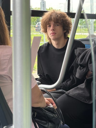 The image depicts an indoor scene, likely on a bus, with a young person with light brown, curly hair facing the camera. They are wearing a black long-sleeved shirt. To the left, a portion of another person's shoulder and arm is visible, wearing a light pink shirt. A black handbag rests on the lap of the person in the pink shirt. In the background, through a window, a blurred outdoor environment with greenery and a dark-colored car is visible, suggesting a suburban or semi-rural setting. The overall lighting is natural, coming from the outside. The image does not provide any specific information about the location within Нефтемаш Снт, Russia, beyond the general context of being on a bus.
