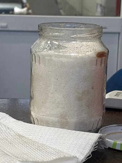 A close-up, eye-level shot shows a glass jar filled with a white granular substance, possibly salt or sugar, sitting on a dark wooden surface. The jar is roughly two-thirds full and has vertical indentations on its exterior. On the right side of the jar, a small, brown object, potentially an insect or piece of debris, is stuck to the glass. In the foreground, a white, textured cloth, possibly a paper towel or napkin, is draped across the surface. A metal lid with green and white stripes is partially visible on the right side of the image, next to the jar. The background is blurred but suggests an indoor environment with light-colored furniture or cabinetry. The scene appears to be located within a city, though no specific landmarks or details indicate the city of Усады, Russia.