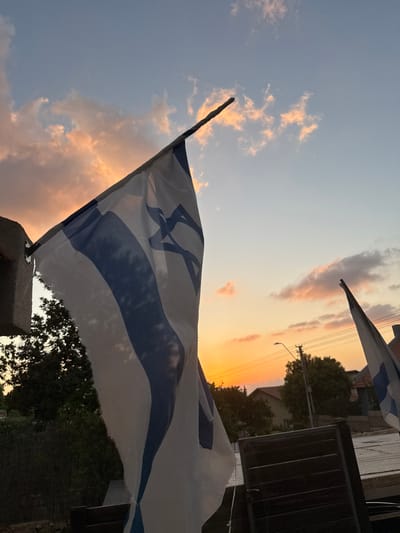 The scene depicts an outdoor environment at either sunrise or sunset, situated in Alfei Menashe, Palestinian Territory. A large Israeli flag, white with two horizontal blue stripes and a blue Star of David, is prominently featured in the left foreground, attached to a light-colored, pillar-like structure. The flag's fabric shows some fraying on its lower edge and is sufficiently translucent to reveal shadows of dense green foliage behind it. A second, smaller Israeli flag is partially visible on the right side of the mid-ground. The sky, occupying the upper portion of the frame, displays a gradient of colors, transitioning from vibrant orange and pink hues near the horizon to lighter blue overhead, with scattered, illuminated clouds. In the background, residential buildings with light-colored roofs, additional green trees, and a utility pole with overhead power lines are visible. A dark, slatted object, possibly a fence or outdoor furniture, is partially visible in the bottom right corner. No individuals or active interactions are present within the frame.