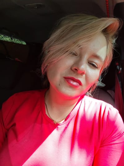A woman with blonde hair is captured in a close-up shot, her face angled slightly downwards. She is wearing a bright red t-shirt and a delicate gold necklace. The lighting suggests she is inside a vehicle, with sunlight casting dramatic shadows across her face and clothing. The background is dark and indistinct, hinting at the interior of a car. The scene is likely located in Vorob'evo, Russia, given the prompt.