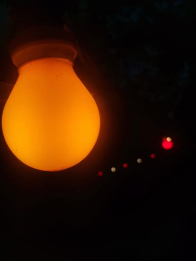 A close-up, low-angle shot focuses on a single, large, orange light bulb hanging in the darkness. The bulb is the dominant feature, emitting a warm glow that illuminates its surface. The background is largely black, with a few out-of-focus, bokeh lights visible in the distance to the right of the bulb. These distant lights appear to be red and white, arranged in a staggered line. The scene suggests an outdoor setting at night, possibly a string of decorative lights. The location in Asprovalta, Greece is not directly depicted but could be inferred as a likely setting for such lighting. There are no people or actions visible in the image.