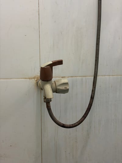 A close-up, eye-level shot shows a weathered, beige plastic faucet attached to a white tiled wall. A brown and beige handle protrudes from the top of the faucet, and a flexible, dark gray metal hose curves down from the faucet's base and trails off to the right. The tiled wall displays signs of age and dirt, with visible grout lines and smudges of grime. The lighting is even, casting no significant shadows. The scene appears to be located in a utilitarian space, possibly a bathroom or utility area. The specific location in Tupah, Indonesia, is not evident from the visual information provided.