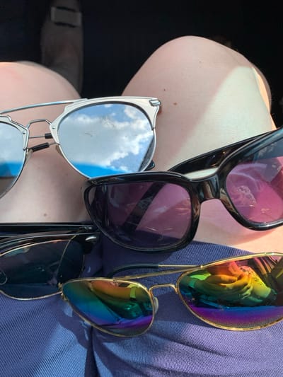 The image shows a close-up, eye-level shot of four pairs of sunglasses resting on blue fabric, likely clothing. The sunglasses are of various styles and colors. The pair closest to the viewer has a gold frame and mirrored lenses reflecting a rainbow of colors. Another pair has a black frame and dark lenses with a pinkish tint. A third pair has a silver frame with mirrored lenses that reflect a blue sky with white clouds. The fourth pair, partially obscured, appears to have a dark frame and dark lenses. The background is out of focus but suggests an indoor setting, possibly a car interior, with hints of a person's legs visible. There is no indication of the location being in Азерскі сельскі Савет, Belarus.