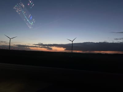 The image depicts an outdoor landscape scene at dusk or early night. The sky transitions from a dark blue-purple at the top to an orange-yellow glow at the horizon, partially obscured by a dark, elongated cloud bank stretching horizontally across the mid-sky. Two three-bladed wind turbines are silhouetted against the horizon; one is positioned on the left, and another further to the right. The foreground is dominated by a dark, undifferentiated landmass. In the distance, beneath the left wind turbine, a horizontal array of small, luminous red and white lights is visible. The upper left quadrant of the image contains a distinct, multicolored reflection consistent with a digital display, exhibiting various lit elements and patterns. This scene is described as being located in or near Châtenay, France.