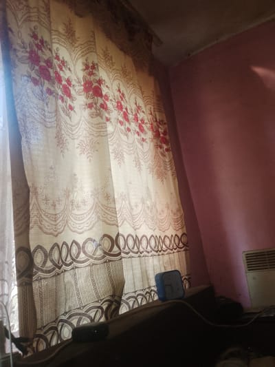 In Fanchykovo, Ukraine, an interior scene is depicted. A patterned curtain, featuring floral motifs in red and beige, dominates the left side of the frame, letting in diffused light from an unseen window. To the right, a pink wall creates a stark contrast. On a dark surface in the foreground, a blue alarm clock displays the time 11:12. A white radiator is visible on the wall in the upper right corner. The scene suggests a domestic environment.