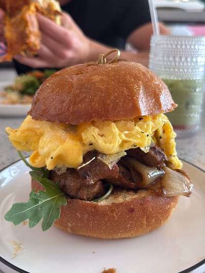 The image shows a breakfast sandwich sitting on a white plate on a textured gray surface. The sandwich consists of a soft bun, arugula, caramelized onions, what appears to be braised short rib, and scrambled eggs. A toothpick or skewer pierces the top bun. In the background, blurred figures of people are visible. One person is holding a piece of food. A textured glass filled with a green liquid is also in the background, along with a plate of food. The setting suggests a restaurant or cafe, likely in Palm Desert, United States.