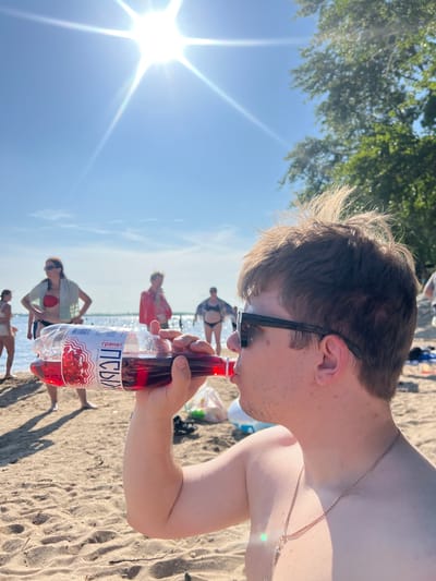 A man is drinking from a plastic bottle of red liquid on a sunny beach in Ust'ye, Russia. He is wearing sunglasses and a chain necklace. In the background, several people are visible near the water and on the sand. The sun is bright in the sky, and there are trees in the distance. The beach is sandy.