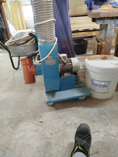 The image depicts a workshop environment in Kaleytsa, Bulgaria. In the foreground, a person's foot wearing a black work boot with green laces is visible, suggesting they are standing on a concrete floor. The floor is littered with sawdust and debris, indicative of woodworking activities.
Dominating the mid-ground is a blue dust collector machine with a corrugated hose attached. To the left of the dust collector, a wheelbarrow filled with wooden planks is situated. A large white bucket with a red lid, labeled "HERMAN VIL DHW/1," stands to the right of the dust collector.
In the background, stacks of lumber are visible, along with a portion of a large blue fabric or curtain. A window can be seen on the far left, letting in natural light. The overall scene suggests an active woodworking or construction site.