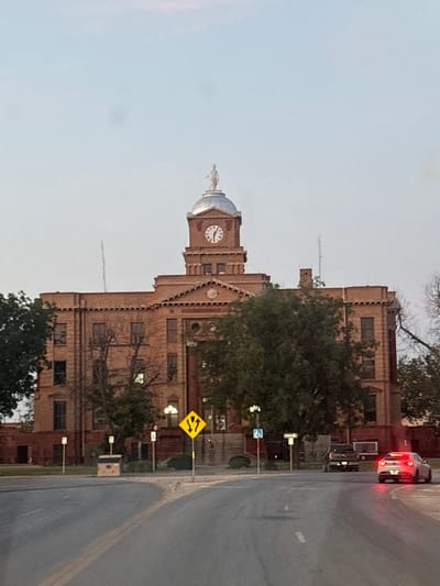 The image depicts a brick courthouse with a domed clock tower in Anson, Texas, USA. The courthouse appears to be old, with visible architectural details like arched windows and decorative stonework. A statue is visible atop the clock tower. The sky is a pale, hazy blue, suggesting either early morning or late evening, with diffused light. The building is partially obscured by trees, some of which are bare, indicating a possible autumn or winter season.

To the right of the courthouse, a street is visible with a car having its brake lights illuminated, suggesting it is stopped or slowing down. Road signs are also present, including a yellow diamond-shaped sign with arrows indicating merging traffic. The overall scene has a quiet, somewhat somber atmosphere due to the lighting and the muted colors. There is no visible text in the image.