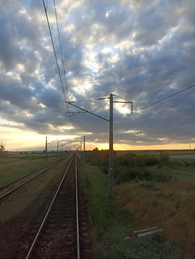 The image depicts a railway line stretching into the distance under a dramatic sky filled with clouds at sunset. The sun is low on the horizon, casting a warm, golden light across the landscape and creating a contrast with the darker, shadowed clouds. Overhead, electrical wires for the train are visible, suspended from poles that line the tracks. The tracks themselves are flanked by grassy fields and low-lying vegetation. The scene is static and tranquil, with no people or moving vehicles present. The location is identified as the outskirts of Zavoy, Bulgaria.
