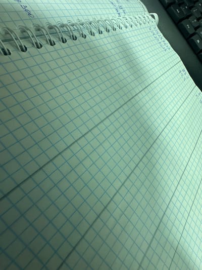 The image shows a close-up, angled view of a notebook with a graph-lined page. The notebook is white with faint blue grid lines. The spine of the notebook, which is white and spiral-bound, is visible along the top left edge. Some handwritten blue ink marks are visible on the page, including what appears to be "p 300" and some numbers such as "31923", "41425", and "933". A portion of a black keyboard is visible in the upper right corner of the image. The lighting suggests an indoor setting. There is no information about the city of Nalazar, Uzbekistan present in the image.