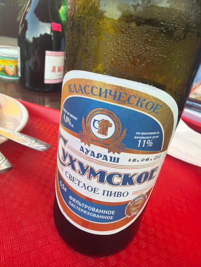 A partially visible, condensation-covered bottle of "Sukhmskoe" light beer is the primary subject of this close-up, eye-level shot. The bottle has a white label with gold and blue accents. Text on the label is in Russian and includes "CLASSIC SKOE," "Sukhmskoe," "LIGHT BEER," and "FILTRATED PASTEURIZED." There is also information about alcohol content (4.0% abv), original gravity (11%), and a date that appears to be "12.06.25." A small circular emblem with a beer mug and "SOCHI" is also visible. The bottle rests on a red textured tray. In the background, out of focus, are parts of other bottles, including a dark bottle with a pink and white label, and a small can with yellow and green markings. A silver fork is visible on the left side of the frame. The lighting suggests an outdoor setting.