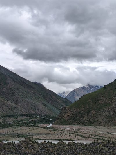 The image shows a wide valley with a river flowing through it. The valley is surrounded by mountains. The sky is overcast with clouds. In the foreground, there are large rocks and sparse vegetation. The middle ground shows the river winding through a dry, rocky landscape dotted with small trees. The background features steep, green mountains on either side of the valley, with snow-capped peaks visible in the distance through gaps in the clouds. The overall impression is one of a remote, mountainous region under a dramatic, cloudy sky. The scene is located in Darcha, India, a high-altitude town in the Lahul and Spiti district of Himachal Pradesh.